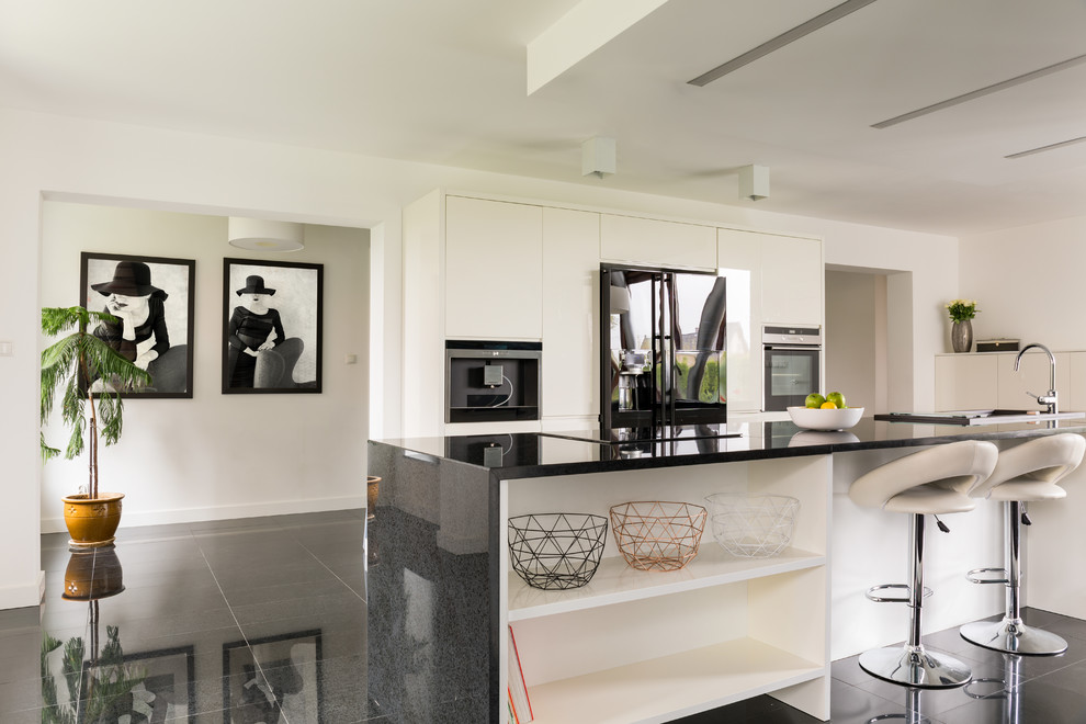 Photo of a contemporary galley kitchen in Los Angeles with a drop-in sink, flat-panel cabinets, white cabinets, black appliances, with island and black floor.