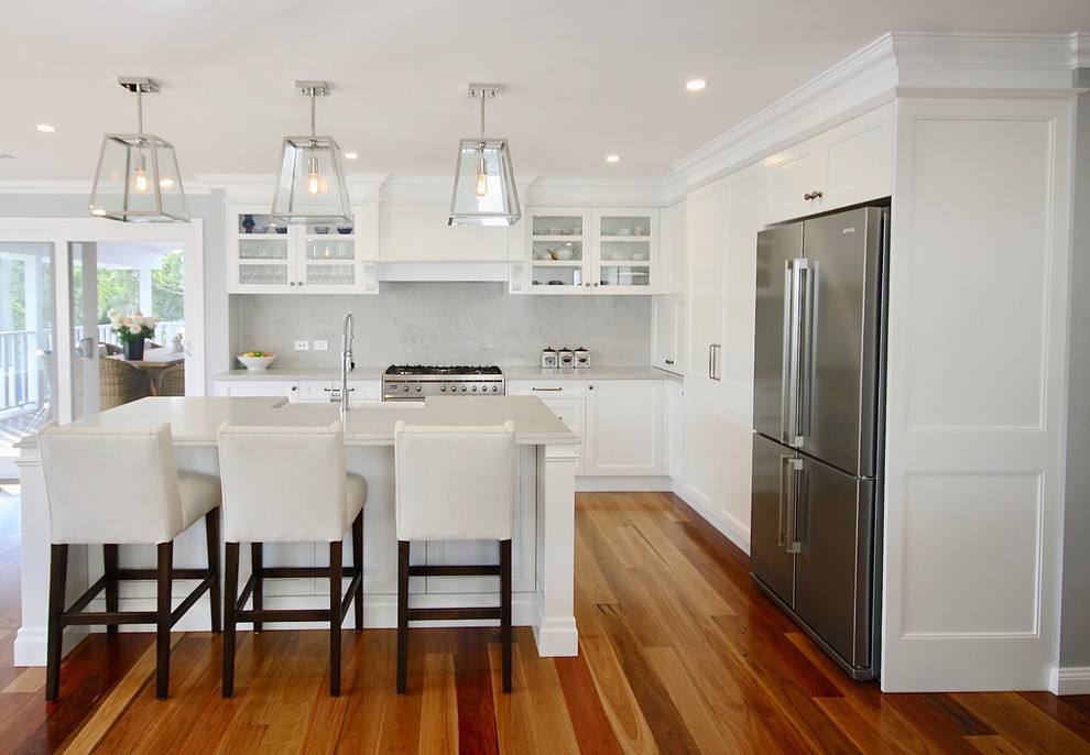 Photo of a large transitional l-shaped eat-in kitchen in Sydney with a farmhouse sink, recessed-panel cabinets, white cabinets, quartz benchtops, grey splashback, stone slab splashback, stainless steel appliances, medium hardwood floors, with island and brown floor.