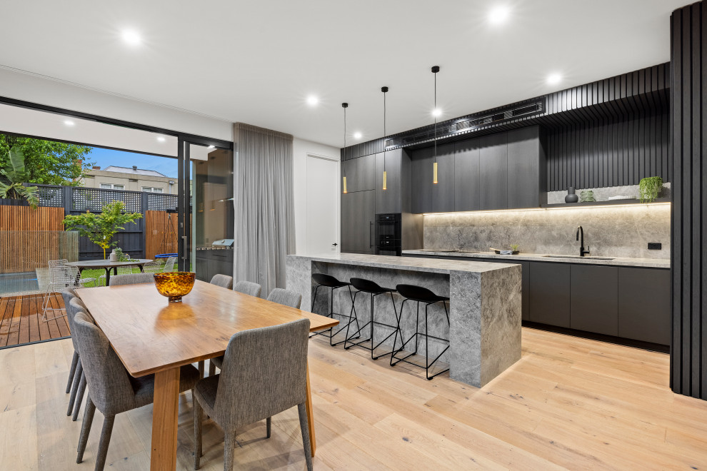 Mid-sized contemporary galley eat-in kitchen in Melbourne with dark wood cabinets, marble benchtops, grey splashback, marble splashback, black appliances, light hardwood floors, with island and grey benchtop.