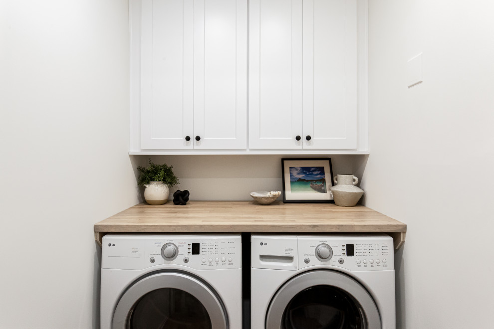 Foto på en liten funkis bruna parallell tvättstuga enbart för tvätt, med skåp i shakerstil, vita skåp, träbänkskiva, vita väggar, ljust trägolv, en tvättmaskin och torktumlare bredvid varandra och beiget golv
