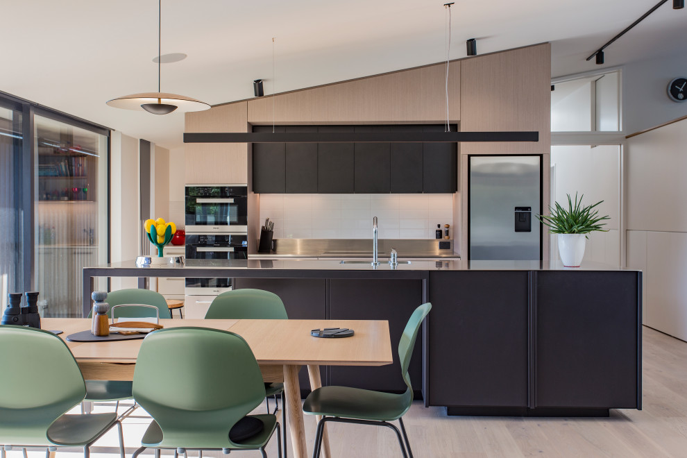 Photo of a mid-sized contemporary galley open plan kitchen in Christchurch with an integrated sink, black cabinets, stainless steel benchtops, white splashback, porcelain splashback, stainless steel appliances, light hardwood floors, with island, beige floor and flat-panel cabinets.