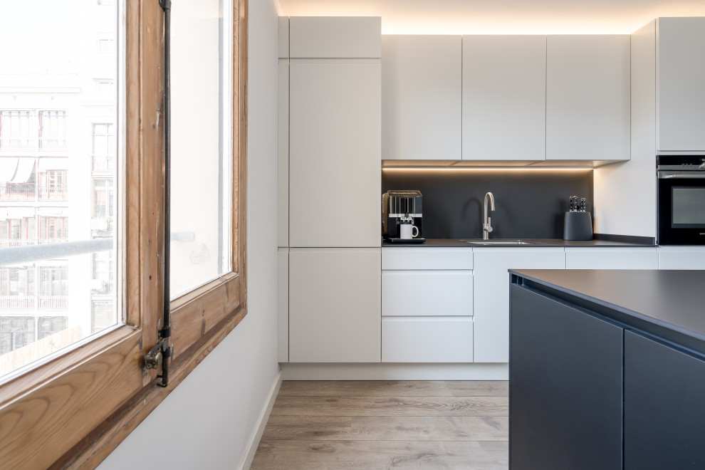 Mid-sized modern u-shaped eat-in kitchen in Other with a single-bowl sink, flat-panel cabinets, white cabinets, limestone benchtops, black splashback, slate splashback, panelled appliances, light hardwood floors, with island, brown floor and black benchtop.