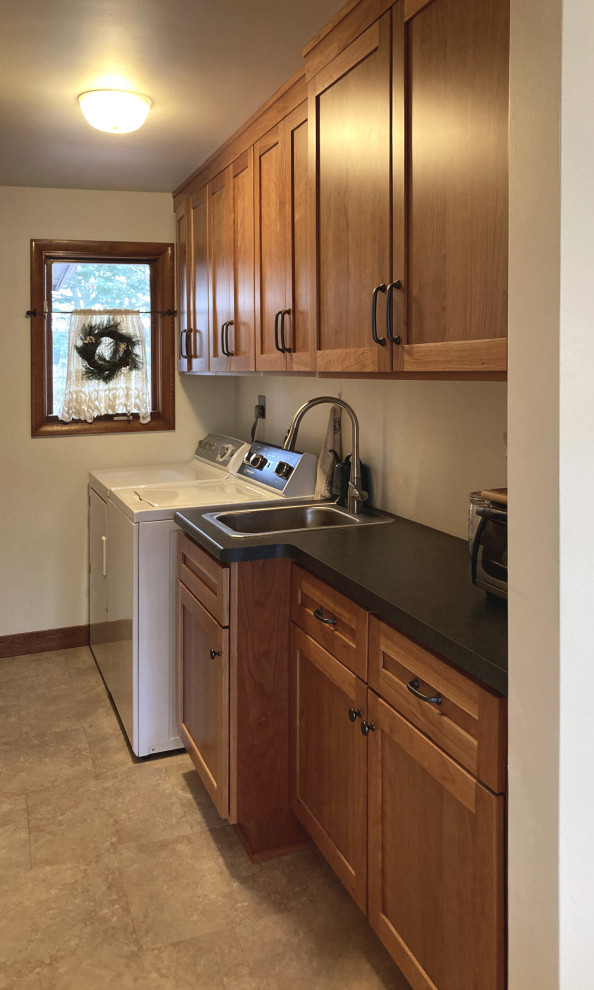 This is an example of a medium sized galley utility room in Other with shaker cabinets, medium wood cabinets, laminate countertops, vinyl flooring, beige floors, black worktops, an utility sink, white walls and a side by side washer and dryer.