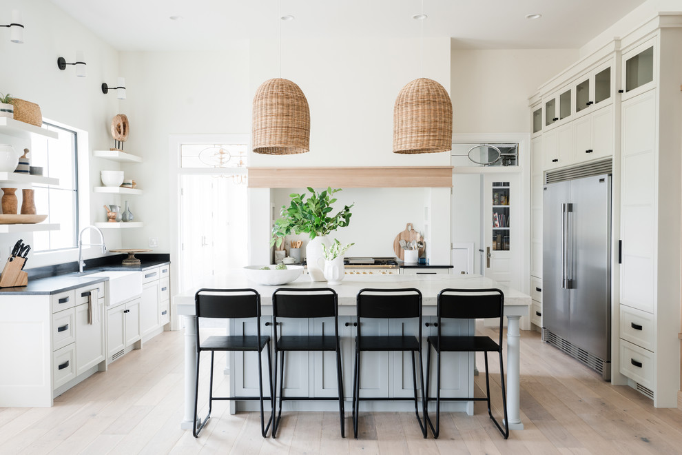 This is an example of a beach style kitchen in Other with a farmhouse sink, recessed-panel cabinets, white cabinets, stainless steel appliances, light hardwood floors, with island, beige floor and black benchtop.