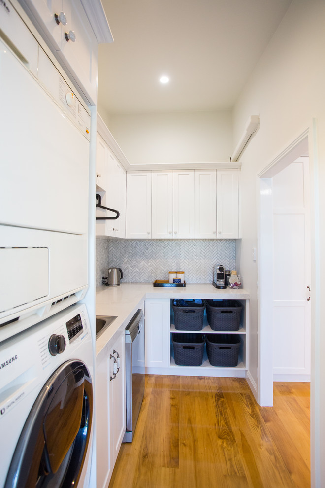 Photo of a small contemporary l-shaped utility room in Brisbane with shaker cabinets, quartz benchtops, grey splashback, marble splashback, medium hardwood floors, brown floor, white benchtop, a single-bowl sink, white cabinets, white walls and a stacked washer and dryer.