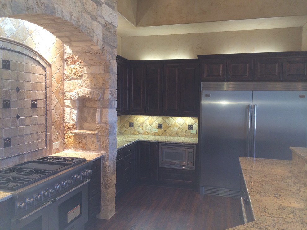 This is an example of a large mediterranean l-shaped eat-in kitchen in Austin with a single-bowl sink, raised-panel cabinets, dark wood cabinets, granite benchtops, beige splashback, stone tile splashback, stainless steel appliances, concrete floors and with island.