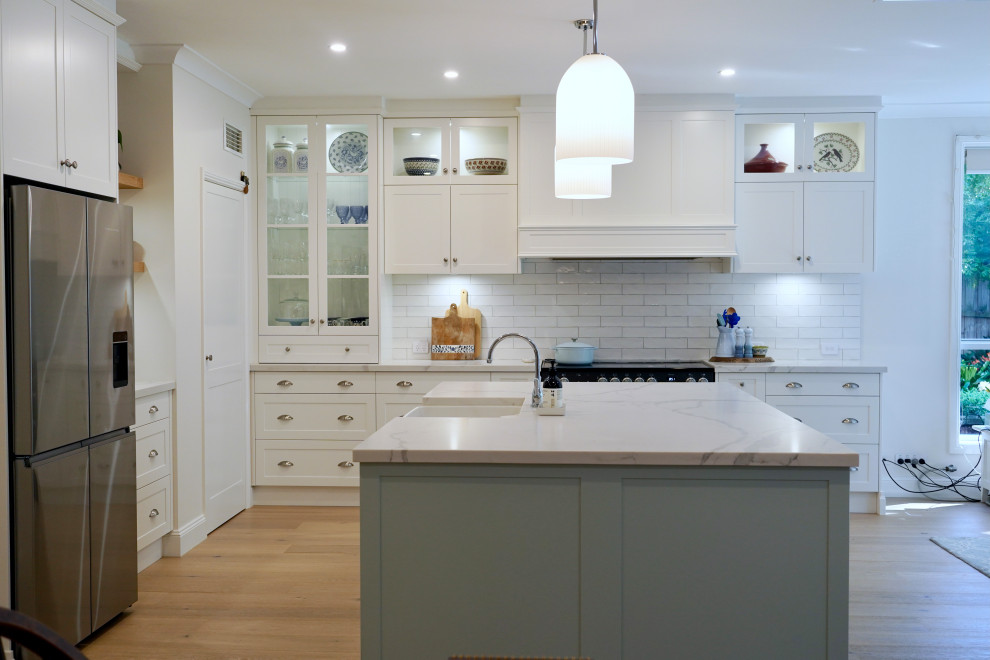 Large traditional l-shaped kitchen/diner in Sydney with a belfast sink, shaker cabinets, white cabinets, engineered stone countertops, white splashback, metro tiled splashback, black appliances, medium hardwood flooring, an island, brown floors and white worktops.