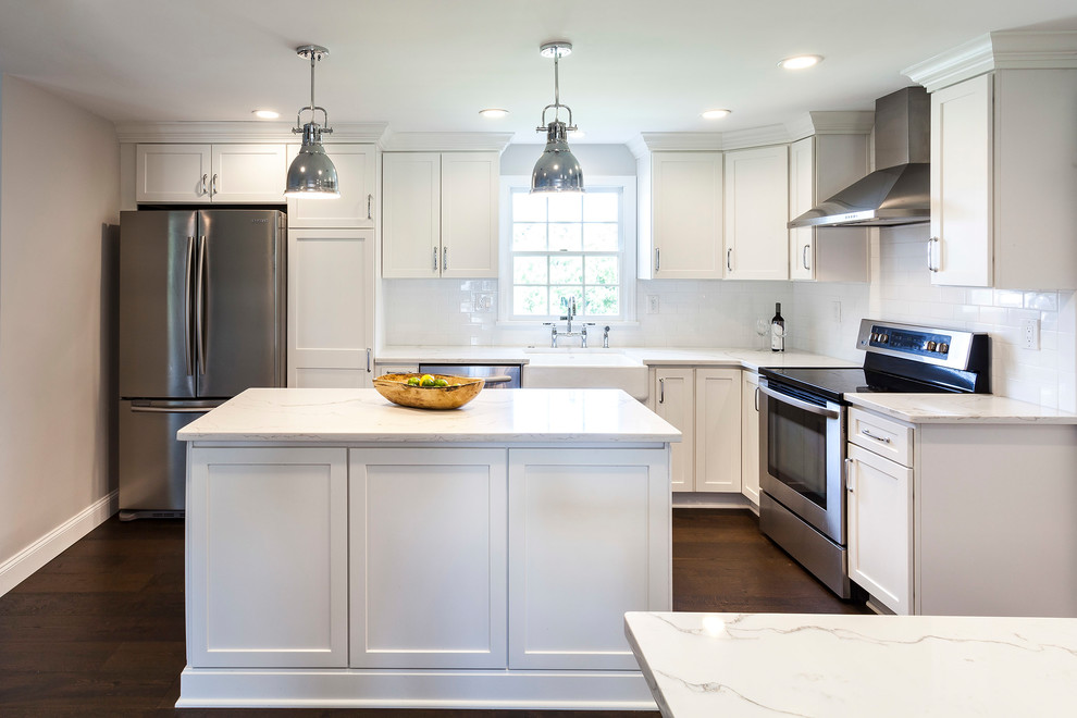 Inspiration for a transitional medium tone wood floor and brown floor kitchen remodel with a farmhouse sink, white cabinets, quartz countertops, white backsplash, ceramic backsplash, stainless steel appliances and an island