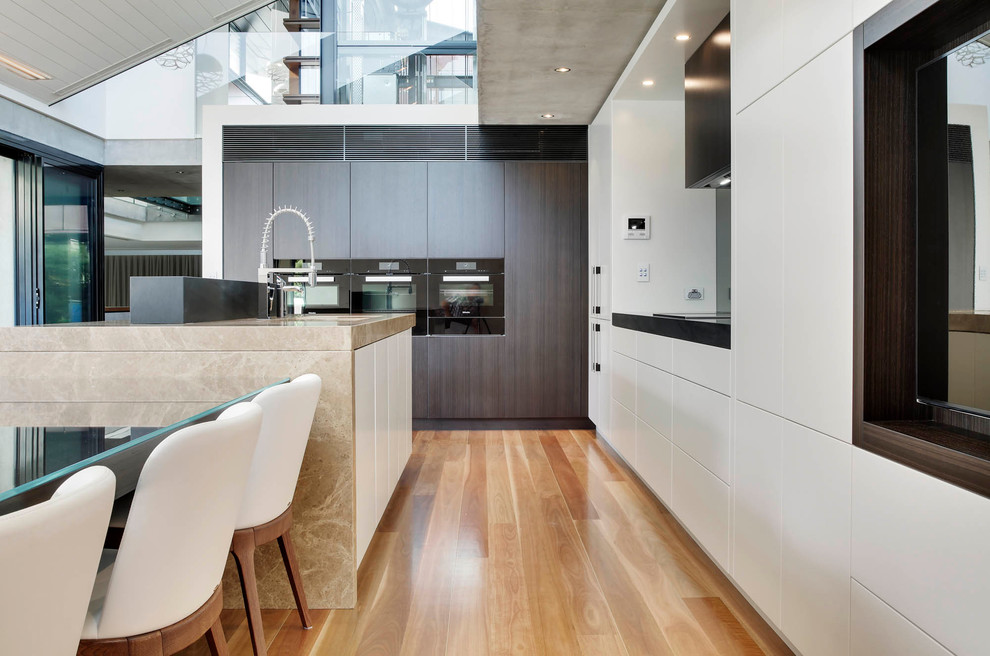 This is an example of an expansive modern l-shaped eat-in kitchen in Sydney with an undermount sink, flat-panel cabinets, beige cabinets, marble benchtops, grey splashback, glass sheet splashback, black appliances, medium hardwood floors and with island.