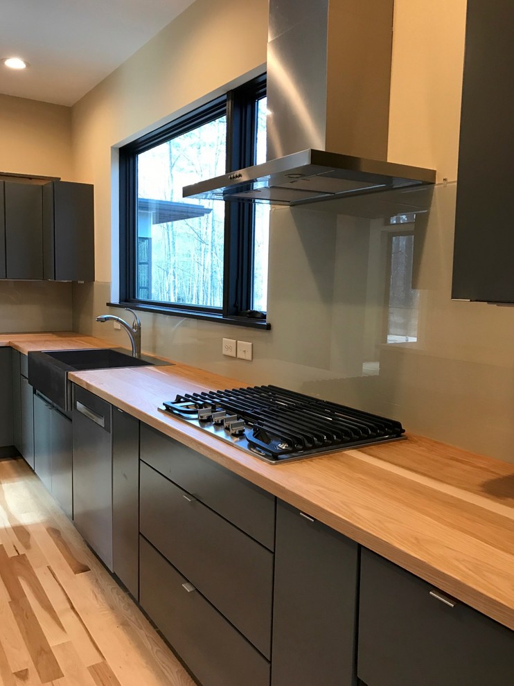 Small modern galley separate kitchen in Raleigh with a farmhouse sink, flat-panel cabinets, grey cabinets, wood benchtops, beige splashback, glass sheet splashback, stainless steel appliances, light hardwood floors and no island.