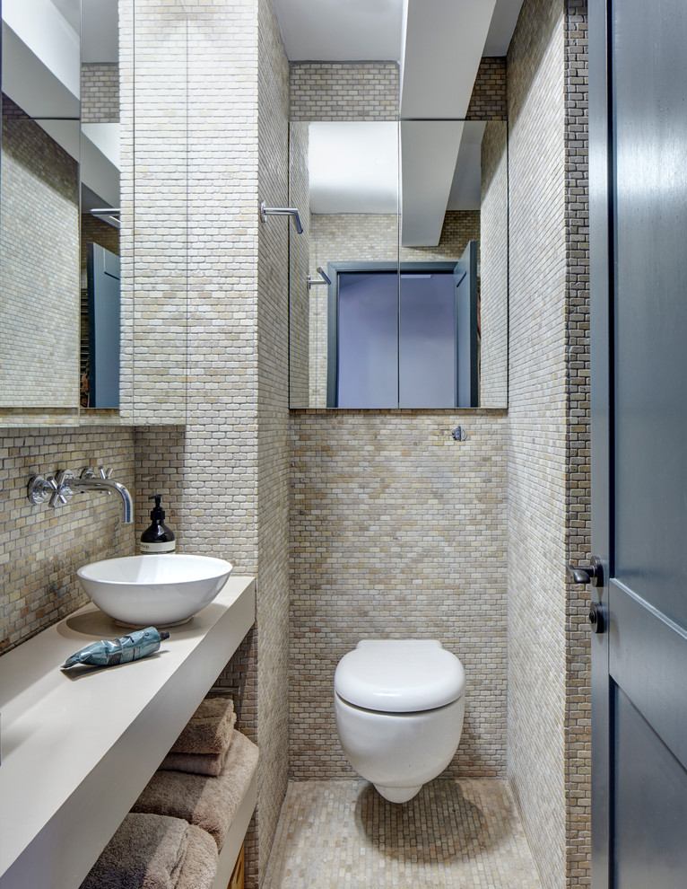 This is an example of a transitional bathroom in London with a vessel sink, open cabinets, a wall-mount toilet and beige tile.