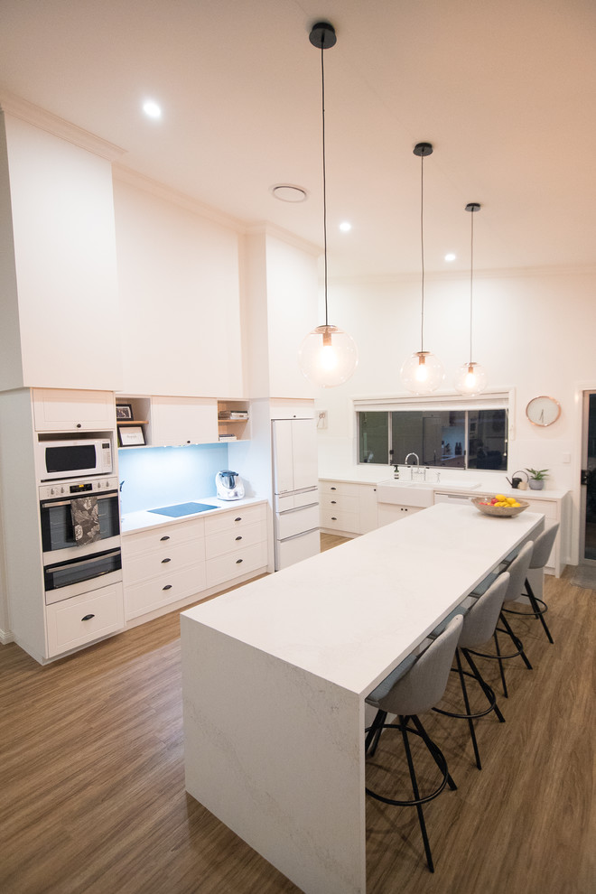 Large modern galley eat-in kitchen in Other with a farmhouse sink, shaker cabinets, white cabinets, marble benchtops, blue splashback, cement tile splashback, white appliances, laminate floors, with island and brown floor.