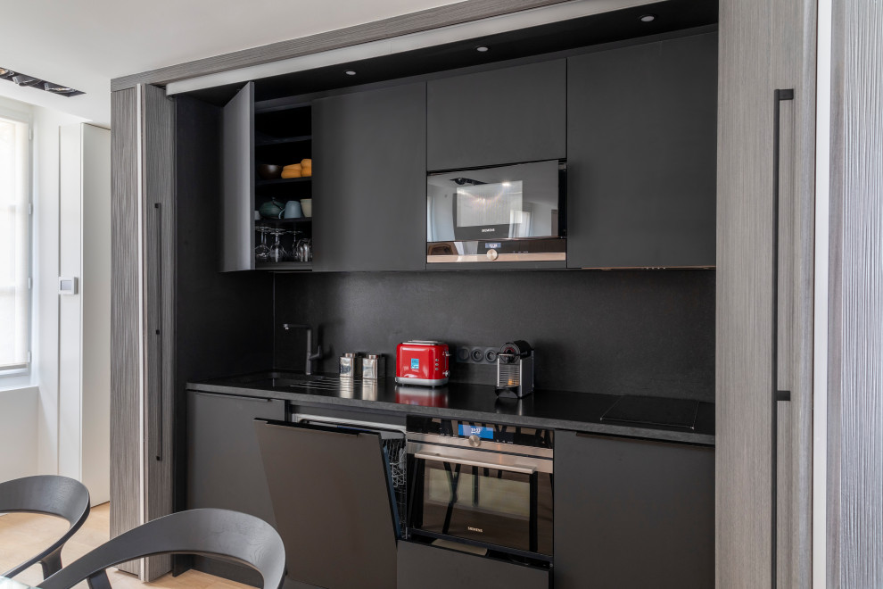 Photo of a small contemporary single-wall open plan kitchen in Other with an undermount sink, beaded inset cabinets, grey cabinets, granite benchtops, black splashback, granite splashback, black appliances, light hardwood floors, no island, beige floor and black benchtop.