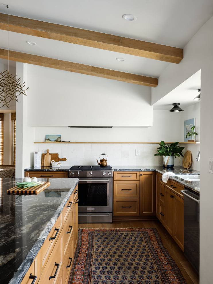 Photo of a mid-sized transitional l-shaped eat-in kitchen in San Diego with an undermount sink, raised-panel cabinets, light wood cabinets, granite benchtops, beige splashback, limestone splashback, stainless steel appliances, light hardwood floors, with island, beige floor, black benchtop and exposed beam.