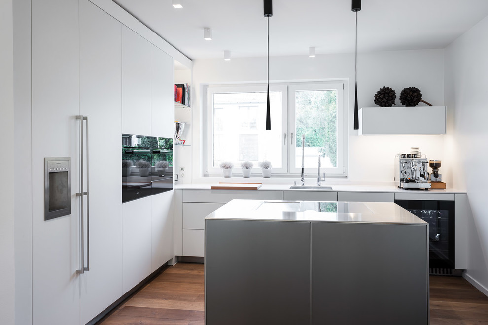 Photo of a mid-sized contemporary l-shaped separate kitchen in Dusseldorf with an integrated sink, flat-panel cabinets, white cabinets, white splashback, black appliances, dark hardwood floors, with island, brown floor and white benchtop.