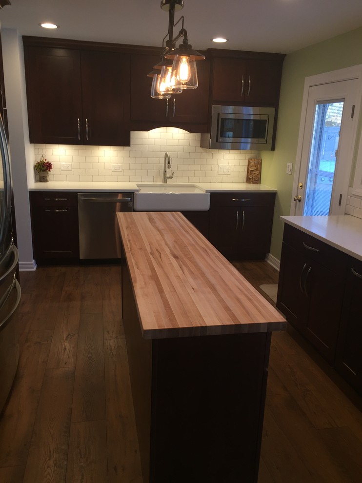 Example of a mid-sized transitional laminate floor and brown floor eat-in kitchen design in Chicago with a farmhouse sink, recessed-panel cabinets, dark wood cabinets, quartz countertops, white backsplash, ceramic backsplash, stainless steel appliances and an island
