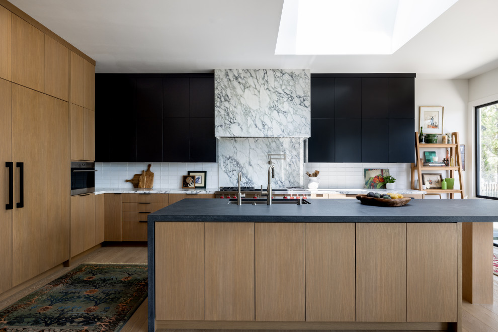 Inspiration for a large contemporary l-shaped eat-in kitchen in Los Angeles with a drop-in sink, flat-panel cabinets, black cabinets, soapstone benchtops, white splashback, marble splashback, stainless steel appliances, medium hardwood floors, with island, brown floor and black benchtop.