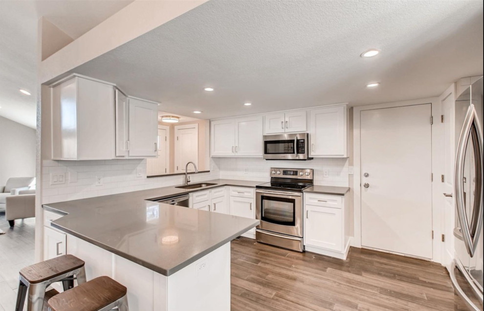 Example of a mid-sized trendy u-shaped medium tone wood floor and brown floor kitchen design with an undermount sink, shaker cabinets, white cabinets, white backsplash, subway tile backsplash, stainless steel appliances, an island and white countertops