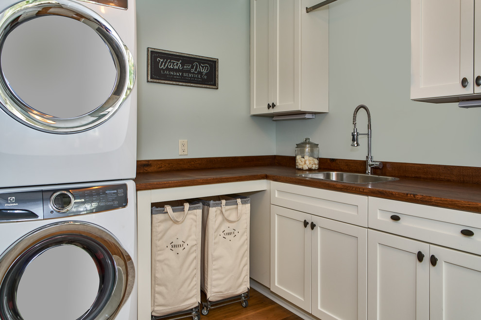 Inspiration for a large beach style l-shaped laundry room in Charleston with a drop-in sink, white cabinets, wood benchtops, medium hardwood floors, a stacked washer and dryer, brown floor, brown benchtop and green walls.