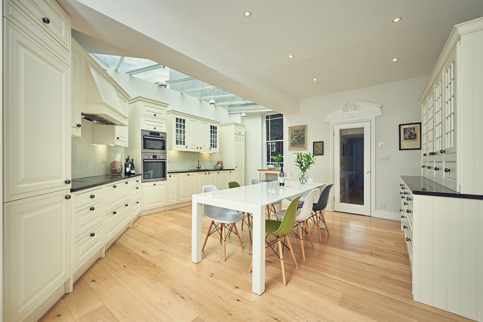 This is an example of a large traditional eat-in kitchen in London with raised-panel cabinets, beige cabinets, beige splashback, glass sheet splashback, light hardwood floors, panelled appliances and no island.