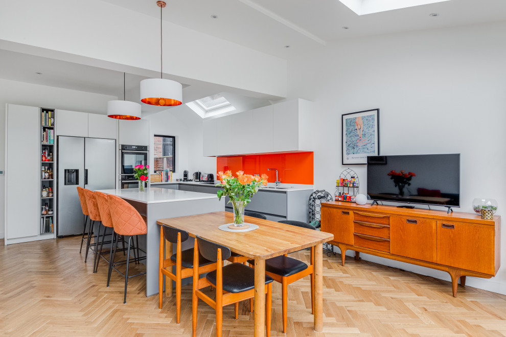 Large contemporary grey and white kitchen/diner in London with a built-in sink, grey cabinets, quartz worktops, orange splashback, glass sheet splashback, coloured appliances, light hardwood flooring, brown floors, white worktops, a vaulted ceiling and feature lighting.
