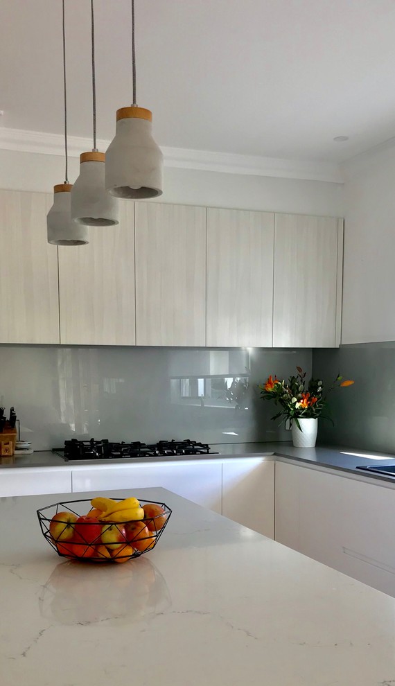 Photo of a mid-sized contemporary l-shaped eat-in kitchen in Melbourne with a double-bowl sink, white cabinets, marble benchtops, metallic splashback, glass sheet splashback, black appliances, light hardwood floors, with island, brown floor and grey benchtop.