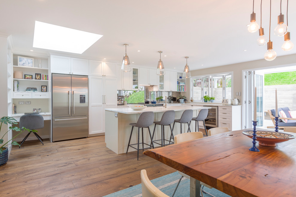Inspiration for a large transitional l-shaped eat-in kitchen in Auckland with an undermount sink, recessed-panel cabinets, white cabinets, quartz benchtops, stainless steel appliances, medium hardwood floors, with island, brown floor, white benchtop and mirror splashback.