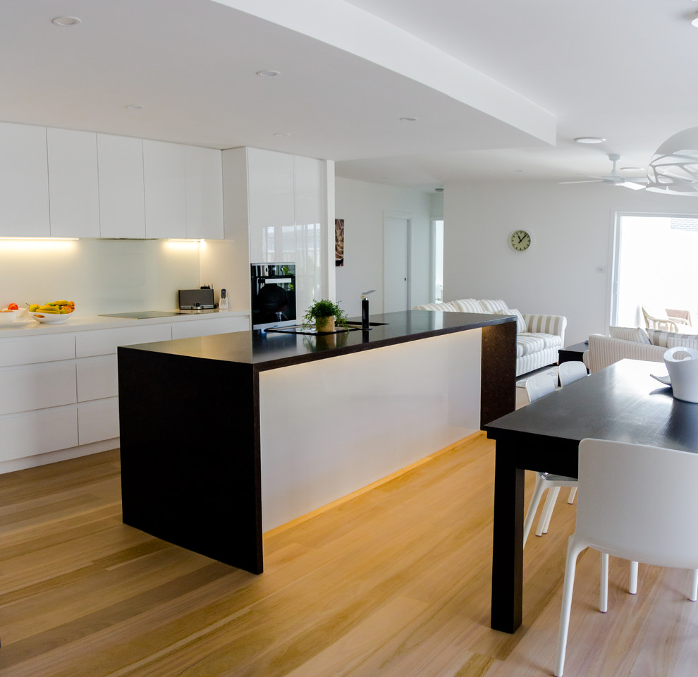 This is an example of a large contemporary l-shaped kitchen pantry in Newcastle - Maitland with an undermount sink, quartz benchtops, white splashback, glass sheet splashback, black appliances, light hardwood floors, with island, flat-panel cabinets and white cabinets.
