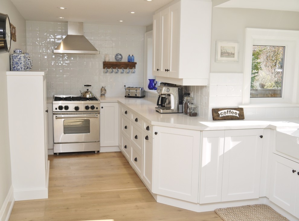 Example of a small transitional l-shaped light wood floor eat-in kitchen design in Toronto with a farmhouse sink, shaker cabinets, white cabinets, quartz countertops, white backsplash, ceramic backsplash, stainless steel appliances and a peninsula
