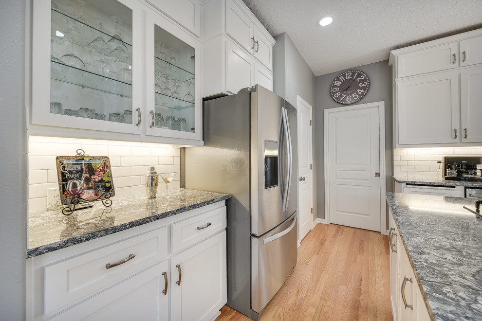 Mid-sized transitional l-shaped light wood floor and brown floor enclosed kitchen photo in Portland with a drop-in sink, shaker cabinets, white cabinets, quartz countertops, white backsplash, subway tile backsplash, an island and stainless steel appliances