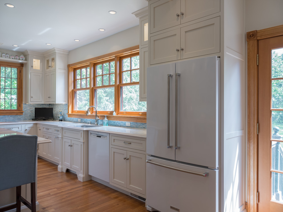 Large traditional l-shaped open plan kitchen in Philadelphia with an undermount sink, flat-panel cabinets, white cabinets, blue splashback, glass tile splashback, white appliances, light hardwood floors, with island and blue floor.
