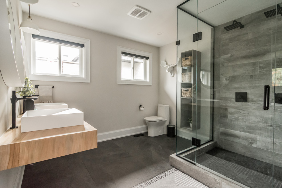 Mid-sized transitional master bathroom in Toronto with open cabinets, brown cabinets, a freestanding tub, a double shower, a one-piece toilet, gray tile, porcelain tile, grey walls, porcelain floors, a vessel sink, wood benchtops, black floor, a hinged shower door and brown benchtops.