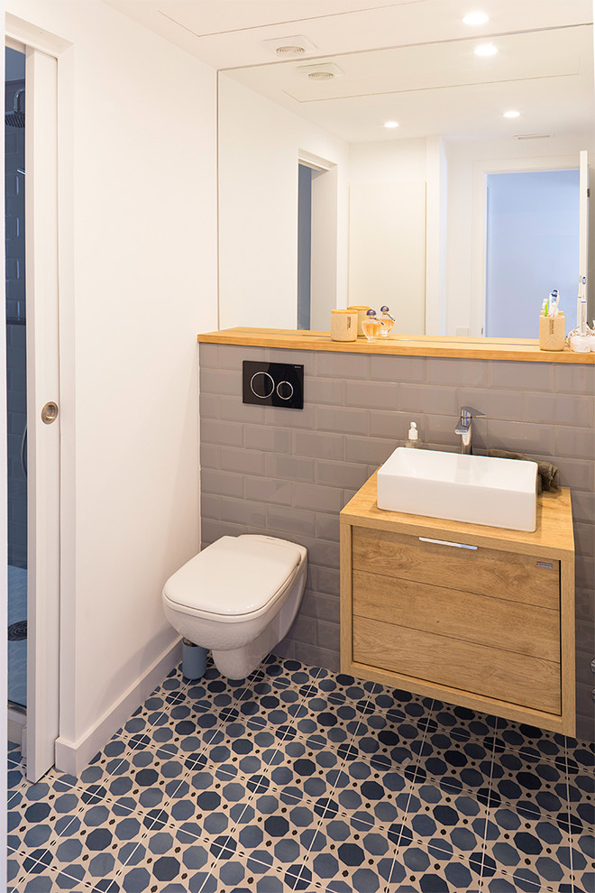 Mid-sized scandinavian bathroom in Barcelona with flat-panel cabinets, light wood cabinets, a wall-mount toilet, gray tile, ceramic tile, white walls, ceramic floors, a vessel sink, wood benchtops, blue floor and brown benchtops.