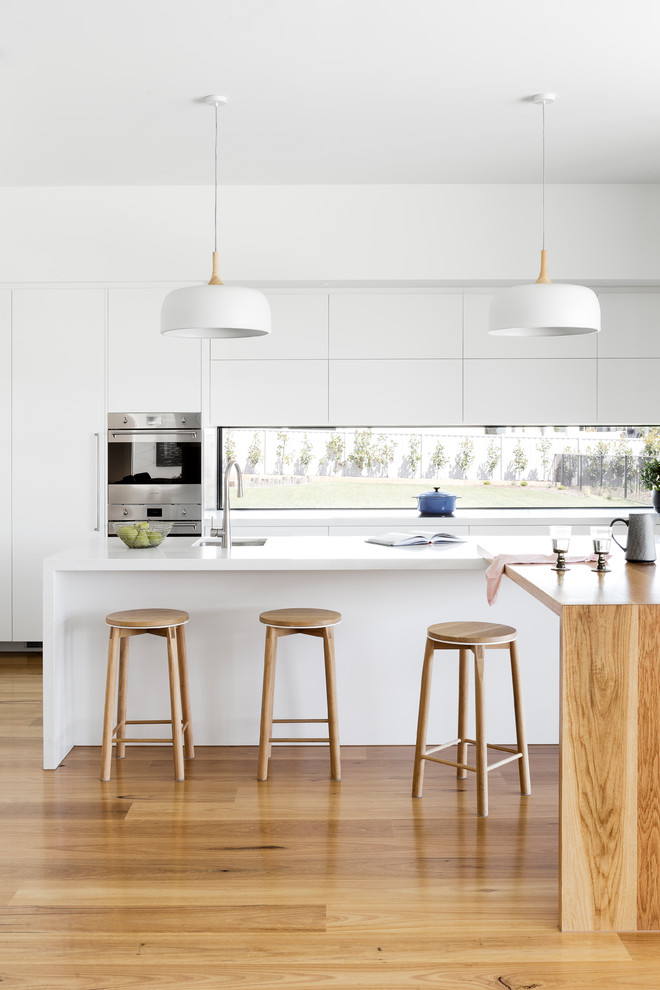 Contemporary galley kitchen pantry in Melbourne with an undermount sink, flat-panel cabinets, white cabinets, quartz benchtops, window splashback, stainless steel appliances, medium hardwood floors, multiple islands, brown floor and white benchtop.