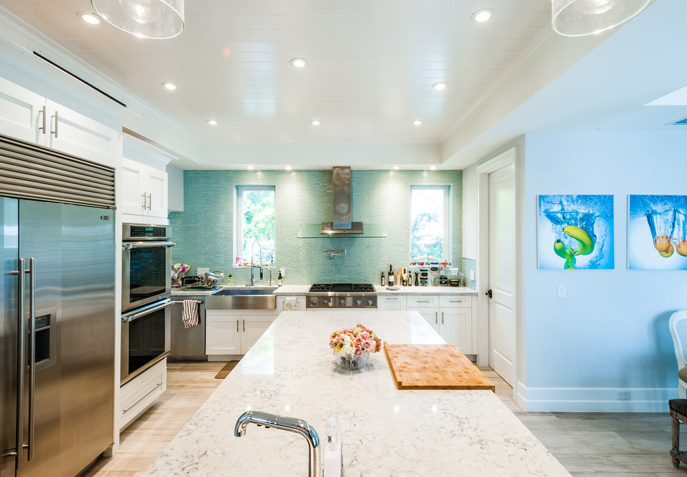 This is an example of a beach style l-shaped open plan kitchen in Miami with a farmhouse sink, shaker cabinets, white cabinets, blue splashback, stainless steel appliances and with island.