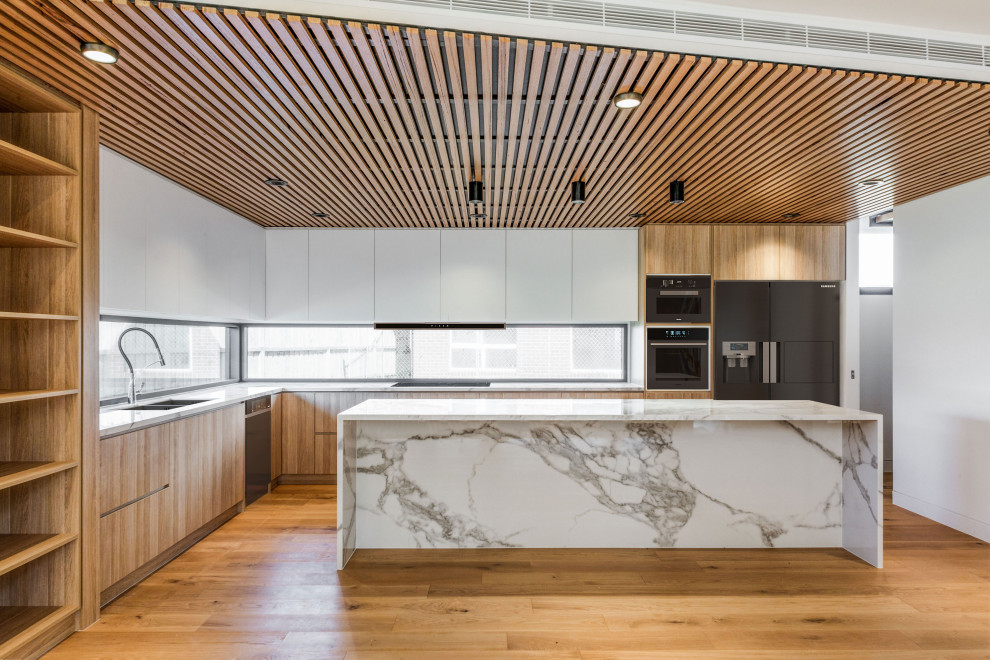This is an example of a large contemporary l-shaped open plan kitchen in Melbourne with a double-bowl sink, light wood cabinets, marble benchtops, window splashback, black appliances, light hardwood floors, with island, white benchtop and wood.