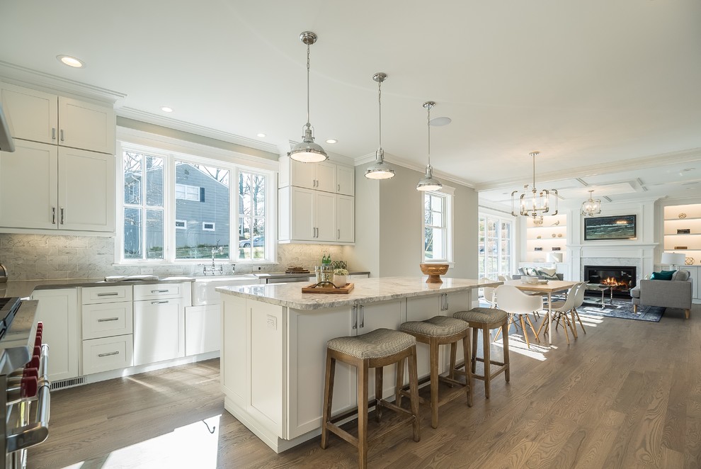 Inspiration for a transitional l-shaped open plan kitchen in New York with a farmhouse sink, shaker cabinets, white cabinets, grey splashback, stainless steel appliances, medium hardwood floors, with island, brown floor and grey benchtop.
