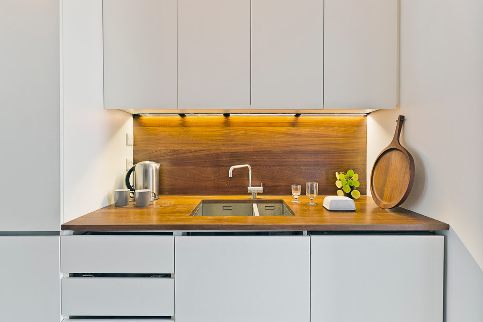 Small midcentury galley separate kitchen in Cambridgeshire with a drop-in sink, flat-panel cabinets, white cabinets, wood benchtops, brown splashback, timber splashback and stainless steel appliances.