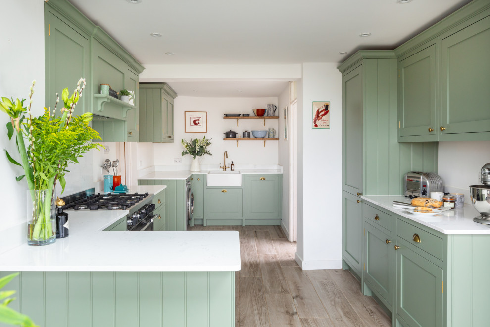 This is an example of a transitional u-shaped kitchen in Other with a farmhouse sink, shaker cabinets, green cabinets, white splashback, panelled appliances, medium hardwood floors, a peninsula, brown floor and white benchtop.