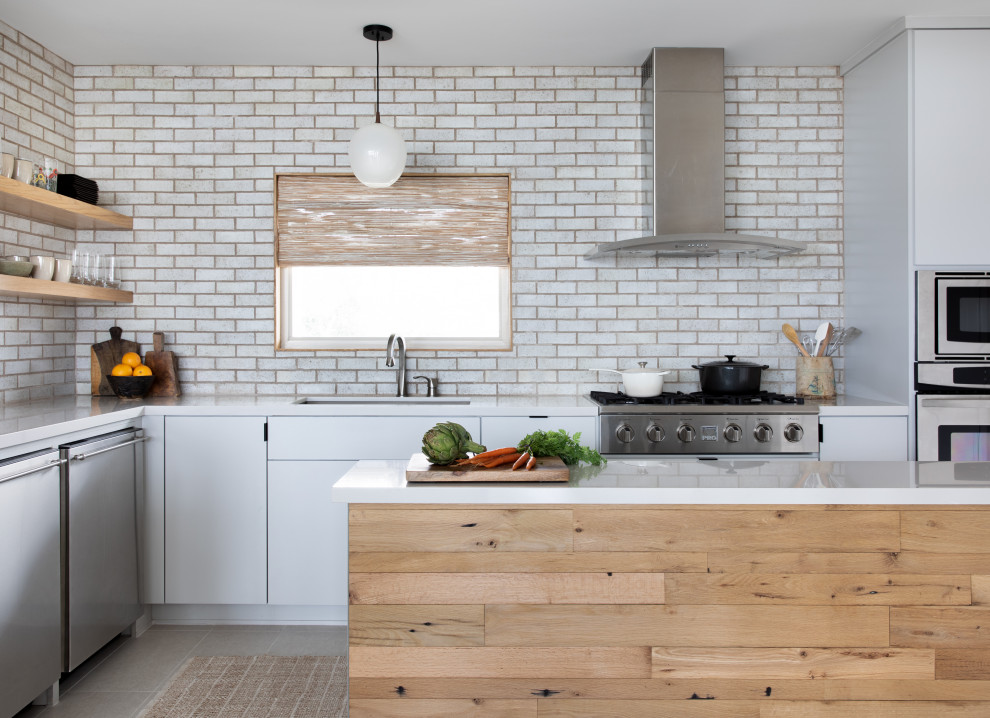 Design ideas for a contemporary l-shaped kitchen with an undermount sink, flat-panel cabinets, white cabinets, white splashback, brick splashback, stainless steel appliances, with island, grey floor and white benchtop.