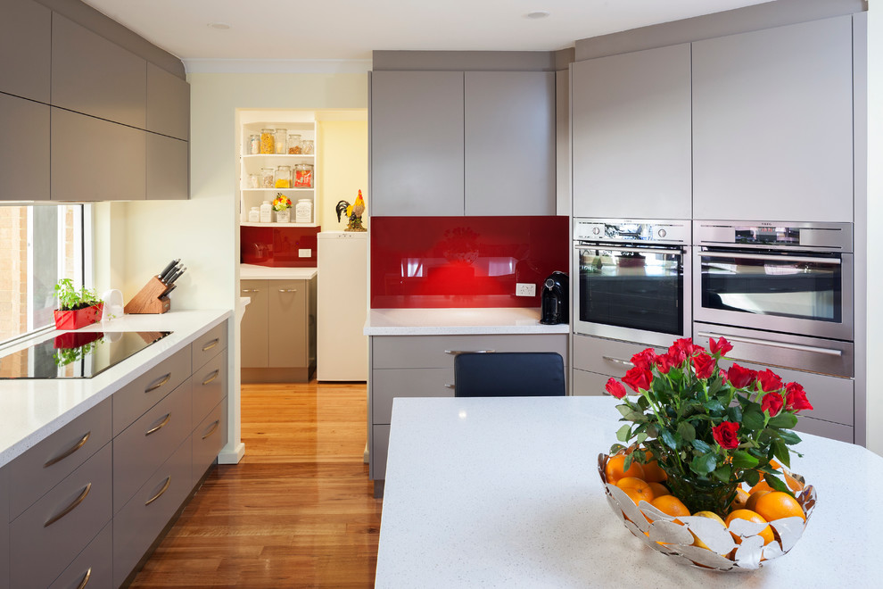 Design ideas for a modern kitchen in Perth with flat-panel cabinets, beige cabinets, quartz benchtops, red splashback, glass sheet splashback, stainless steel appliances, with island and white benchtop.