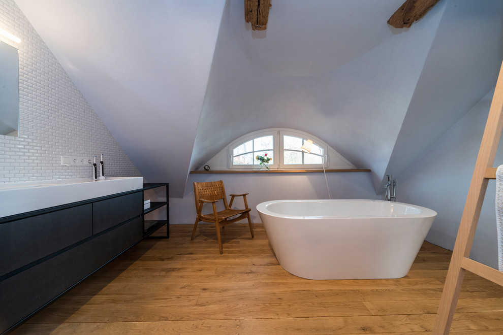 Mittelgroßes Skandinavisches Badezimmer En Suite mit flächenbündigen Schrankfronten, schwarzen Schränken, freistehender Badewanne, weißen Fliesen, Metrofliesen, grauer Wandfarbe, braunem Holzboden, Aufsatzwaschbecken und braunem Boden in Sonstige