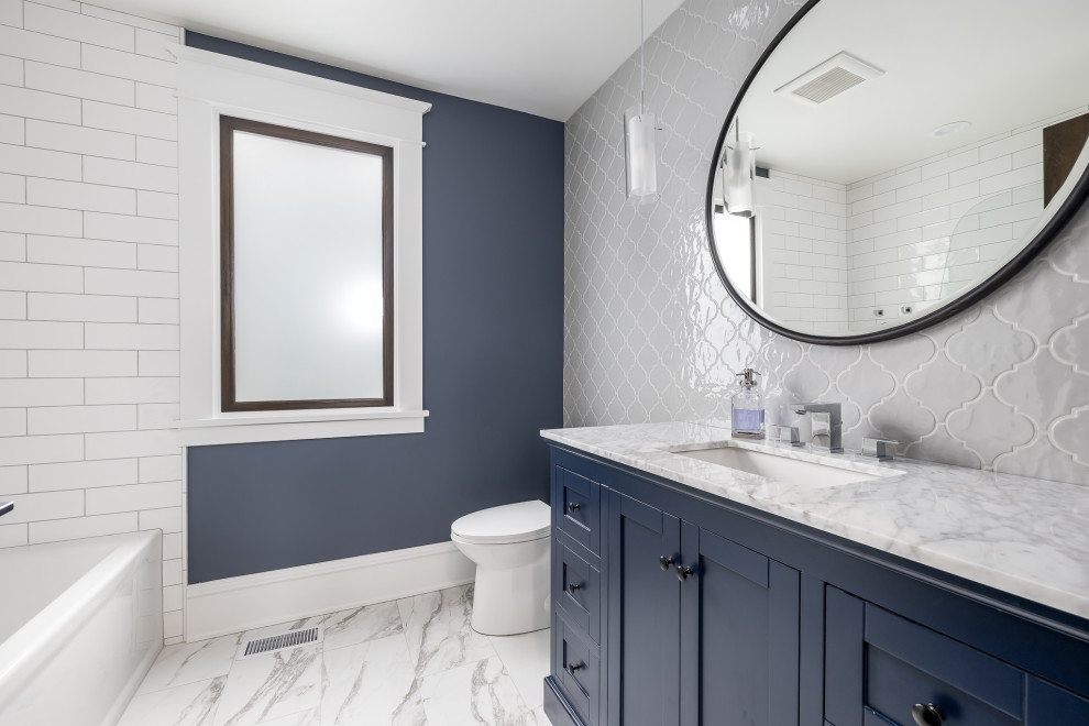 Inspiration for a mid-sized transitional white tile ceramic tile, white floor, single-sink and wainscoting alcove bathtub remodel in Minneapolis with shaker cabinets, blue cabinets, a one-piece toilet, blue walls, an undermount sink, marble countertops, a hinged shower door and a freestanding vanity