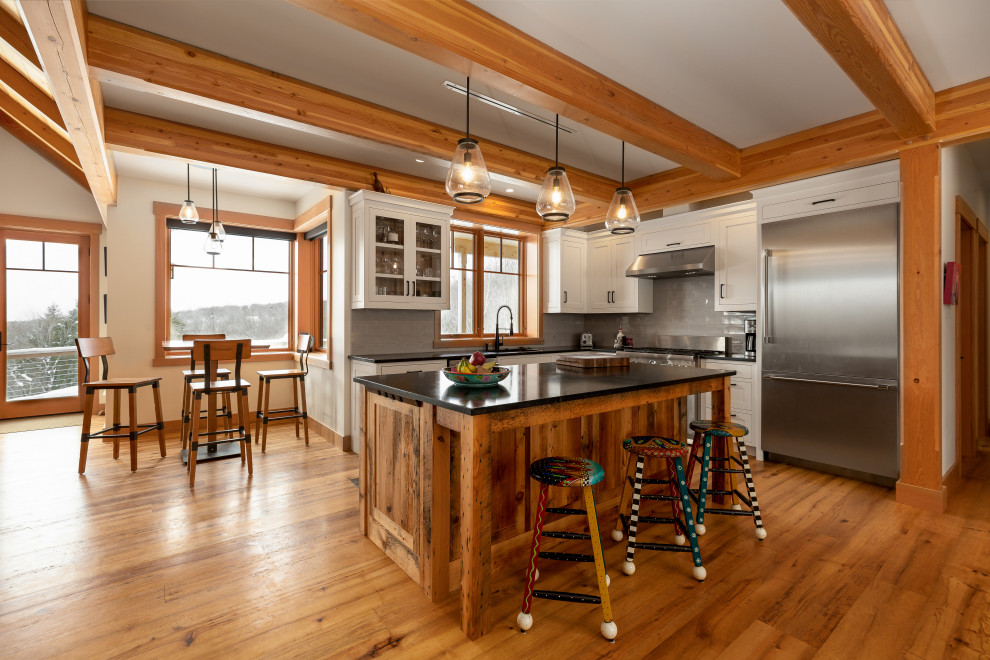 This is an example of a country u-shaped open plan kitchen in Burlington with an undermount sink, shaker cabinets, white cabinets, quartz benchtops, grey splashback, glass tile splashback, stainless steel appliances, medium hardwood floors, with island, brown floor, black benchtop, exposed beam and vaulted.