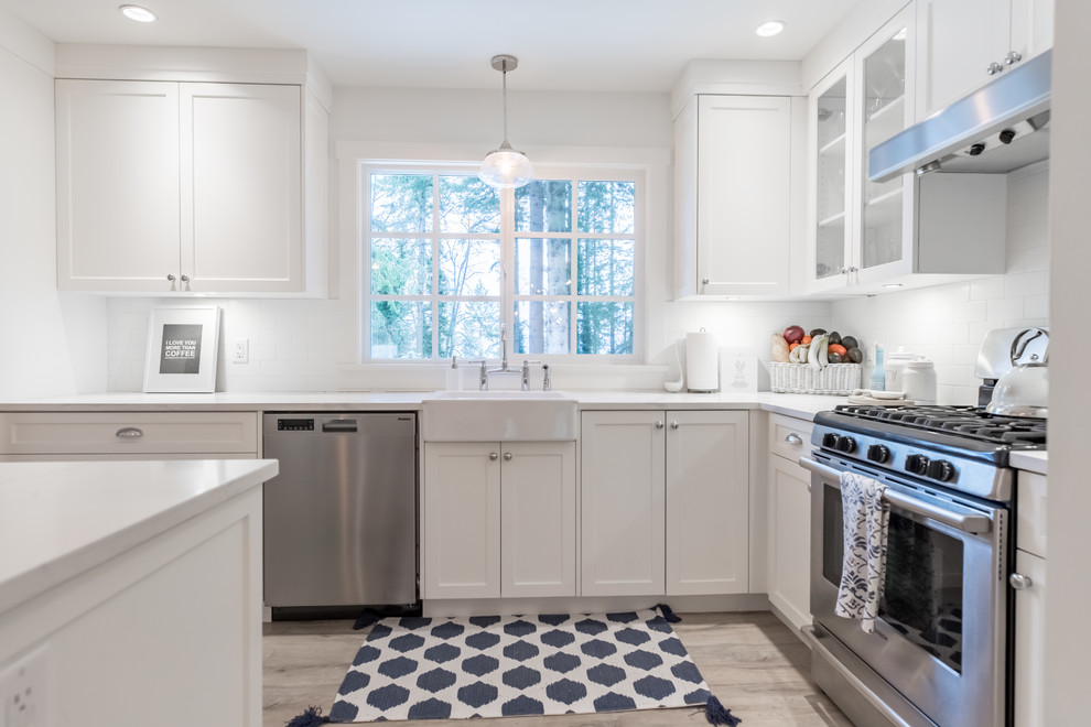Design ideas for a mid-sized arts and crafts u-shaped open plan kitchen in Vancouver with a farmhouse sink, shaker cabinets, white cabinets, quartz benchtops, white splashback, ceramic splashback, stainless steel appliances, laminate floors, with island, beige floor and white benchtop.