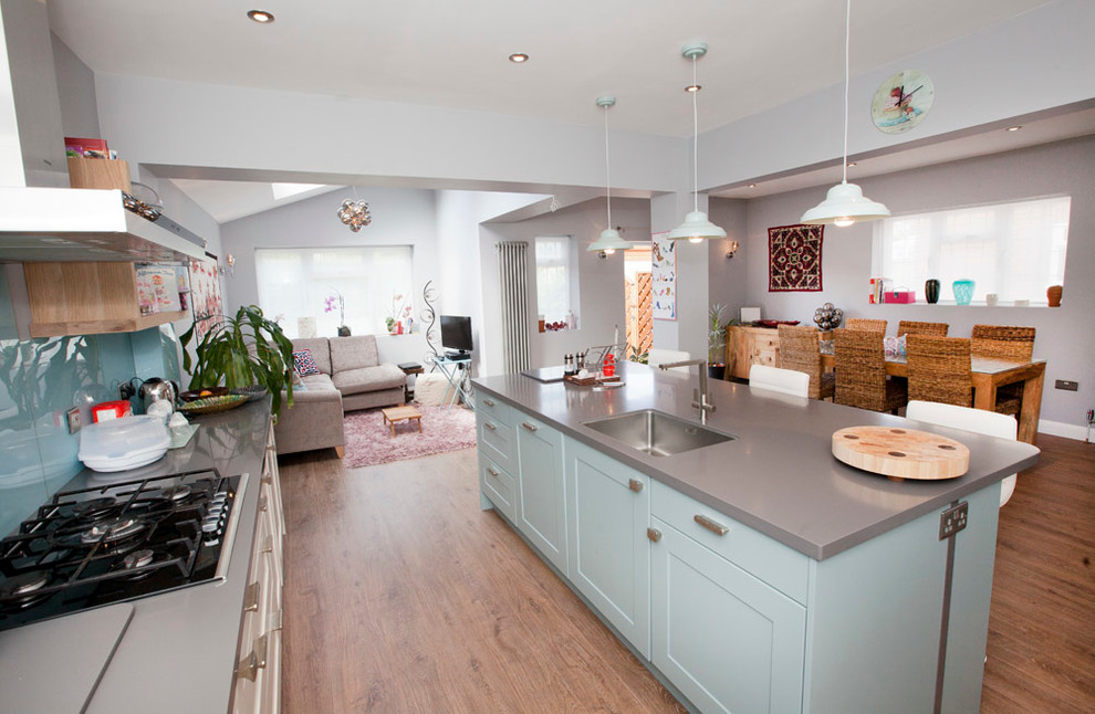 This is an example of a mid-sized eclectic l-shaped open plan kitchen in Other with a drop-in sink, recessed-panel cabinets, white cabinets, solid surface benchtops, blue splashback, glass sheet splashback, stainless steel appliances, medium hardwood floors and with island.
