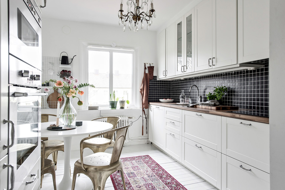 Scandinavian eat-in kitchen in Gothenburg with shaker cabinets, white cabinets, wood benchtops, black splashback, painted wood floors, no island and white floor.