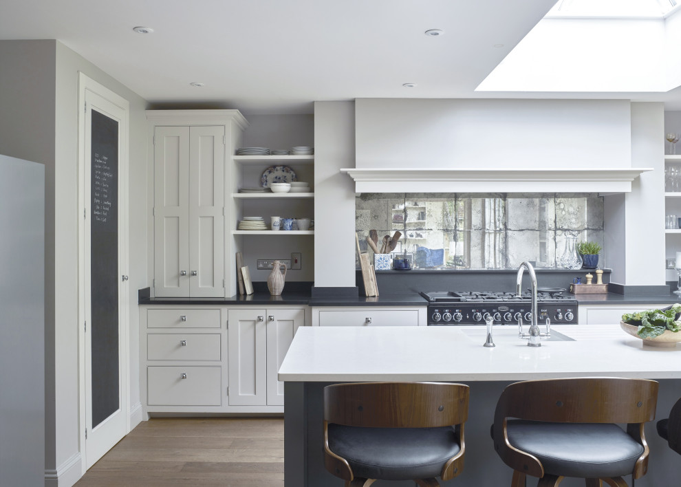 Inspiration for a mid-sized transitional l-shaped open plan kitchen in Surrey with a single-bowl sink, shaker cabinets, white cabinets, granite benchtops, mirror splashback, black appliances, light hardwood floors, with island, beige floor and black benchtop.