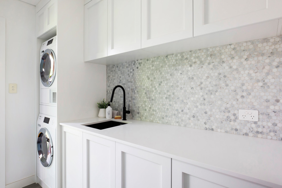 This is an example of a mid-sized beach style galley dedicated laundry room in Sydney with an undermount sink, shaker cabinets, white cabinets, quartz benchtops, grey splashback, mosaic tile splashback, white walls, porcelain floors, a stacked washer and dryer, grey floor, white benchtop, coffered and panelled walls.