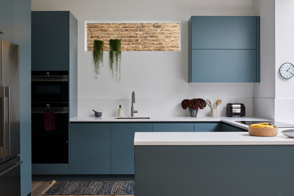 This is an example of a contemporary u-shaped open plan kitchen in London with flat-panel cabinets, blue cabinets, solid surface benchtops, white splashback, engineered quartz splashback, medium hardwood floors, white benchtop, an undermount sink, black appliances, a peninsula and brown floor.